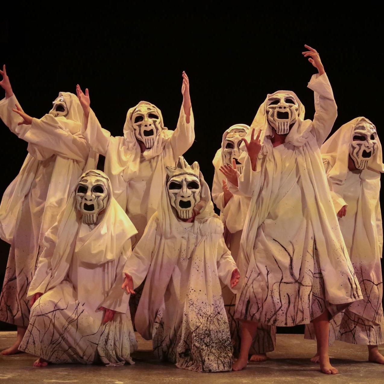 希腊悲剧《底比斯的葬礼》中的合唱队正在舞台上表演. 天黑了. 合唱团成员都穿着白色的破布，戴着白色的口罩. 他们举起手臂，摆出哀悼的姿势.